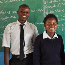 Zambian boy and girl in smiling in front of classroom.