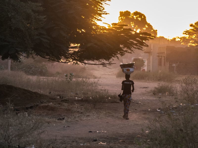 A woman walks down a dirt road carrying goods atop her head in Mali.