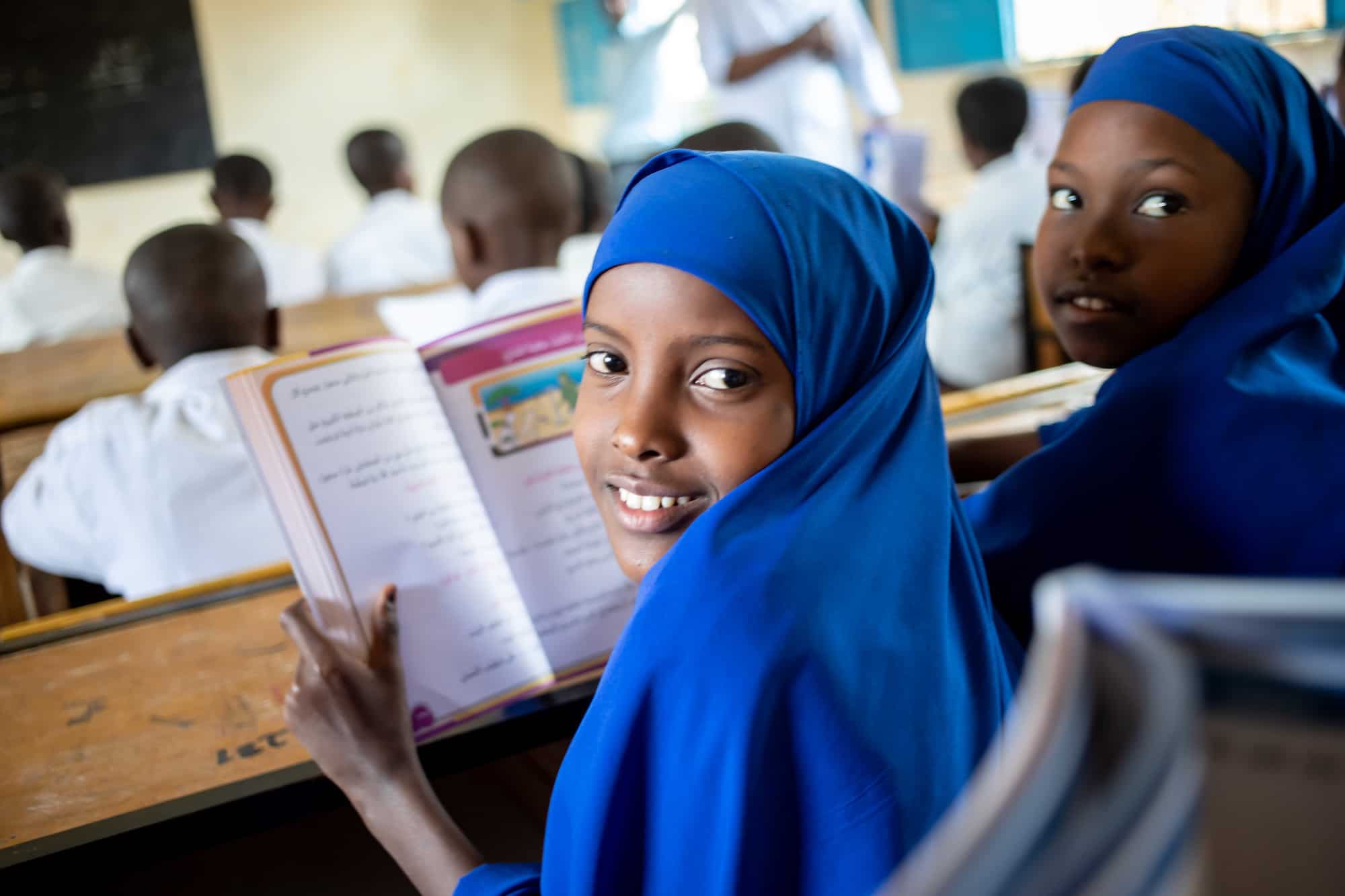 A Somali girl in a blue headscarf sits in the classroom smiling and holding an open book