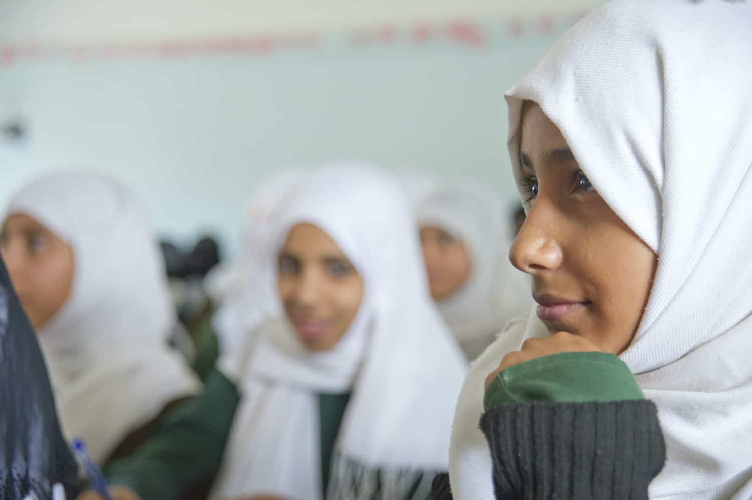 A girl in Yemen wearing a head scarf sits in class, smiling and looking attentive.